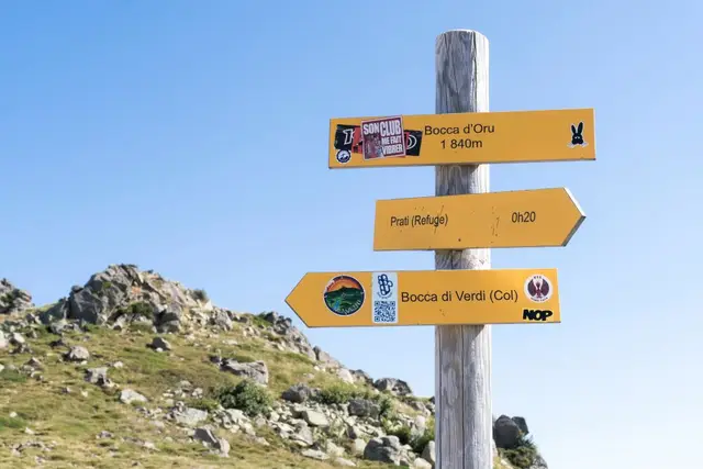 Un panneau de randonnée jaune indique différentes directions et temps de marche en montagne sous un ciel bleu dégagé.